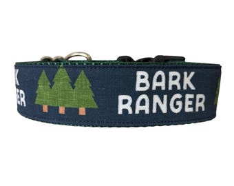 Könnte beinhalten: Ein marineblaues Hundehalsband mit den Worten "BARK RANGER" in weißen Blockbuchstaben. Das Halsband zeigt grüne Kiefern mit braunen Stämmen. Das Halsband hat einen Metallverschluss und eine schwarze Kunststoffschnalle.