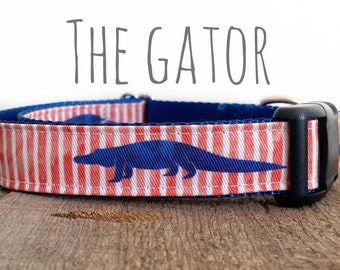 May include: A blue and white striped dog collar with a blue alligator silhouette. The collar has a black buckle and the text "THE GATOR" is printed above the collar.