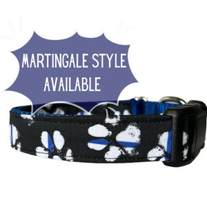 May include: Black dog collar with a white paw print pattern and a thin blue line across each paw print. The collar has a black buckle and a blue trim. The text "MARTINGALE STYLE AVAILABLE" is in a speech bubble above the collar.