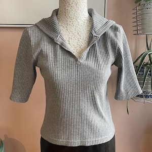 以下が含まれることがあります： グレーのリブニットトップス、ジッパー付きの襟と半袖。トップスは黒のレザースカートと組み合わせています。服はマネキンに展示されています。