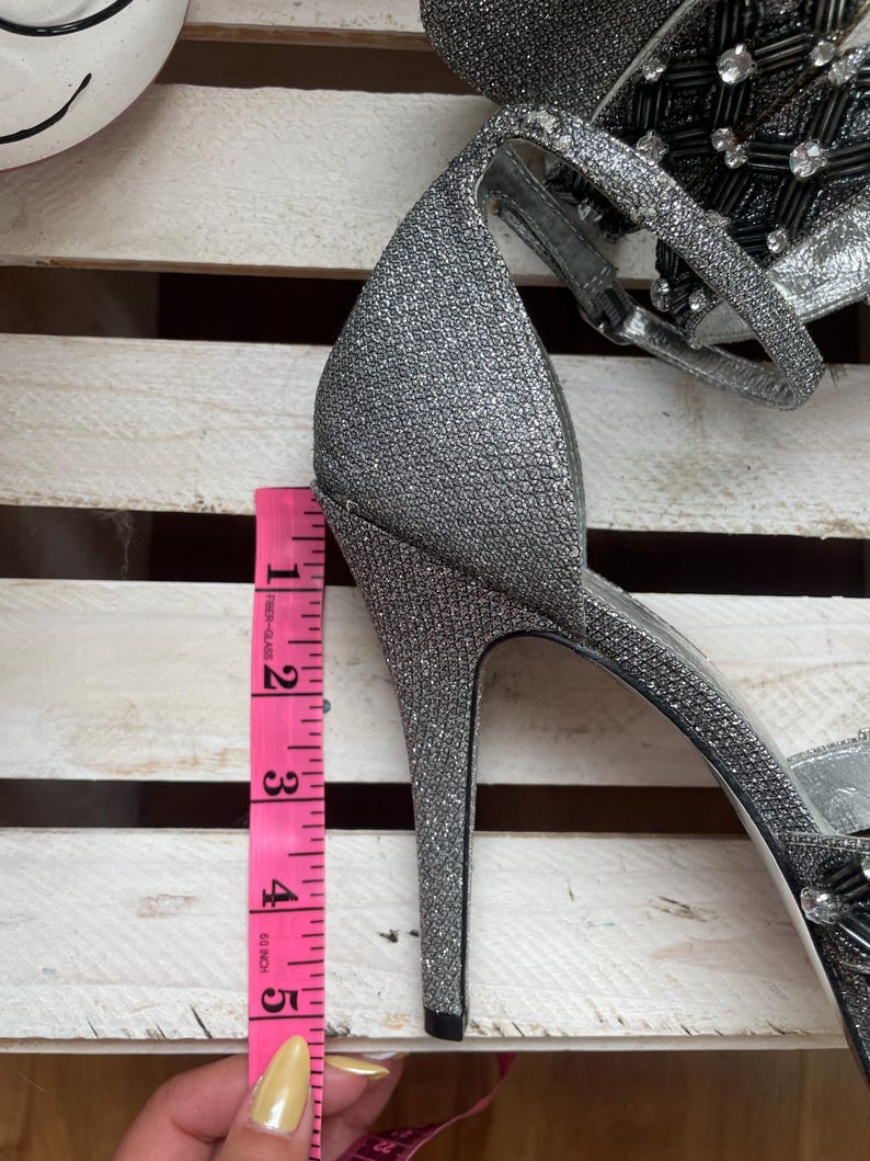 May include: Silver glitter high-heeled sandal with a stiletto heel. The heel measures approximately 4 inches. The sandal features a delicate ankle strap and embellishments on the upper. The shoe is set against a white wooden background.