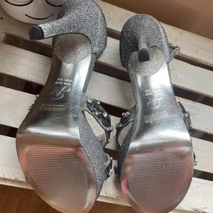 May include: A pair of silver glitter high-heeled sandals. The heels are approximately 3.5 inches tall. The sandals have a strap design with decorative embellishments. The soles are silver and have a textured pattern. The size is USA 9.5M, EUR 39.