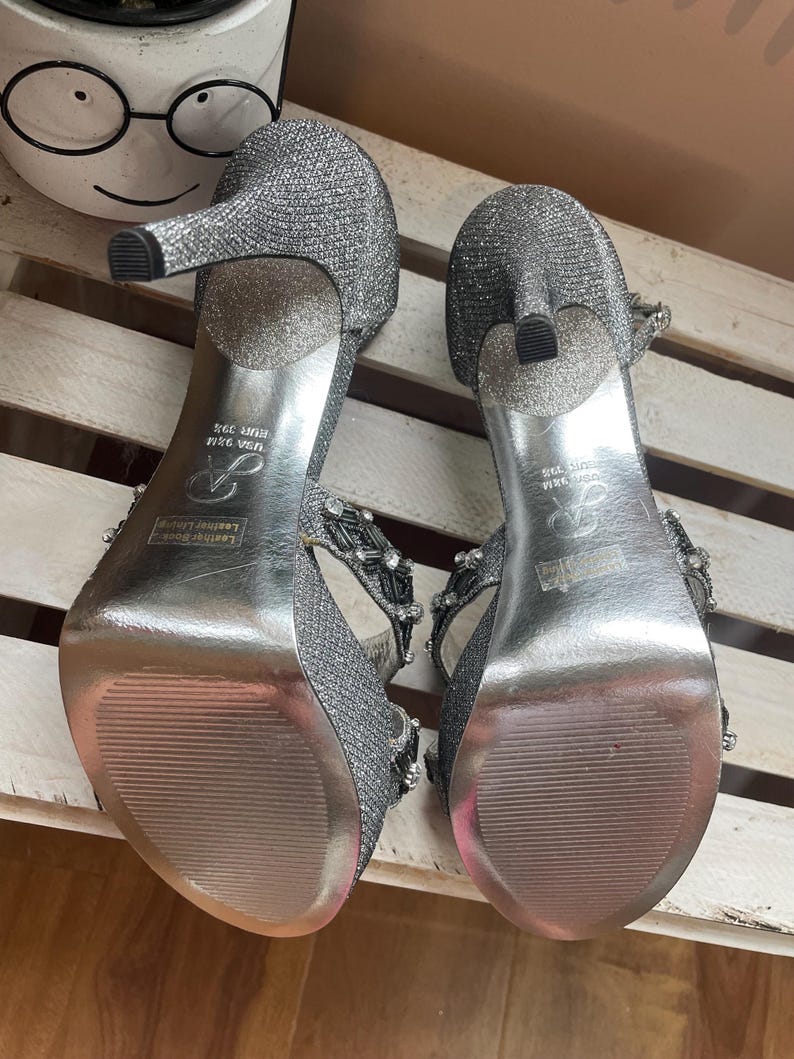 May include: A pair of silver glitter high-heeled sandals. The heels are approximately 3.5 inches tall. The sandals have a strap design with decorative embellishments. The soles are silver and have a textured pattern. The size is USA 9.5M, EUR 39.