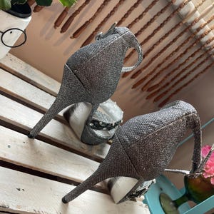 May include: A pair of silver glitter high-heeled shoes with a pointed toe and a strap around the ankle. The heels are slender and the shoes have a textured appearance. The shoes are displayed on a white wooden surface.