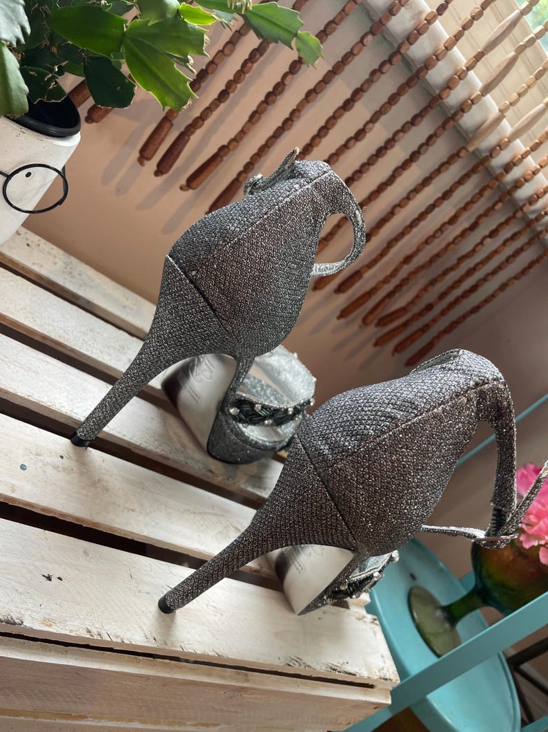 May include: A pair of silver glitter high-heeled shoes with a pointed toe and a strap around the ankle. The heels are slender and the shoes have a textured appearance. The shoes are displayed on a white wooden surface.