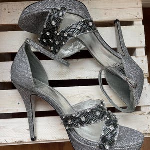 May include: A pair of silver glitter high-heeled sandals with a platform sole. The sandals feature a strap around the ankle and a decorative band across the foot, embellished with black and clear beads. The heels are approximately 4 inches (10 cm) high.