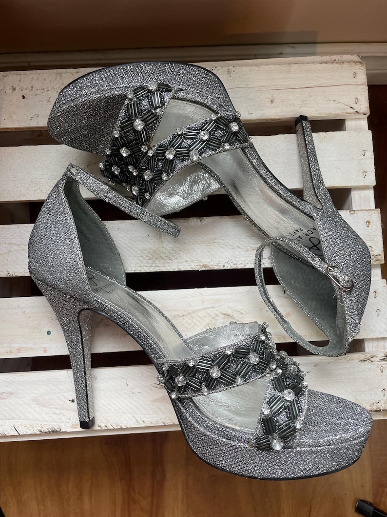 May include: A pair of silver glitter high-heeled sandals with a platform sole. The sandals feature a strap around the ankle and a decorative band across the foot, embellished with black and clear beads. The heels are approximately 4 inches (10 cm) high.
