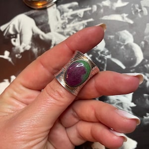 May include: A silver ring featuring a green and pink oval gemstone set in the centre. The ring has a textured band.