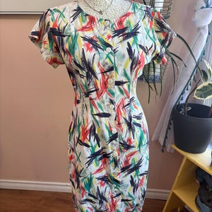 May include: A vintage button-down dress with a colorful abstract brushstroke pattern in red, green, yellow, and black on a white background. The dress has short sleeves and is displayed on a mannequin.