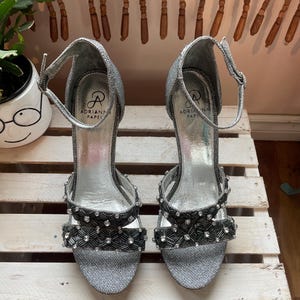 May include: A pair of silver glitter high-heeled sandals with ankle straps. The sandals feature black and silver beaded embellishments on the straps and toe. The brand name "Adrianna Papell" is visible on the insole.