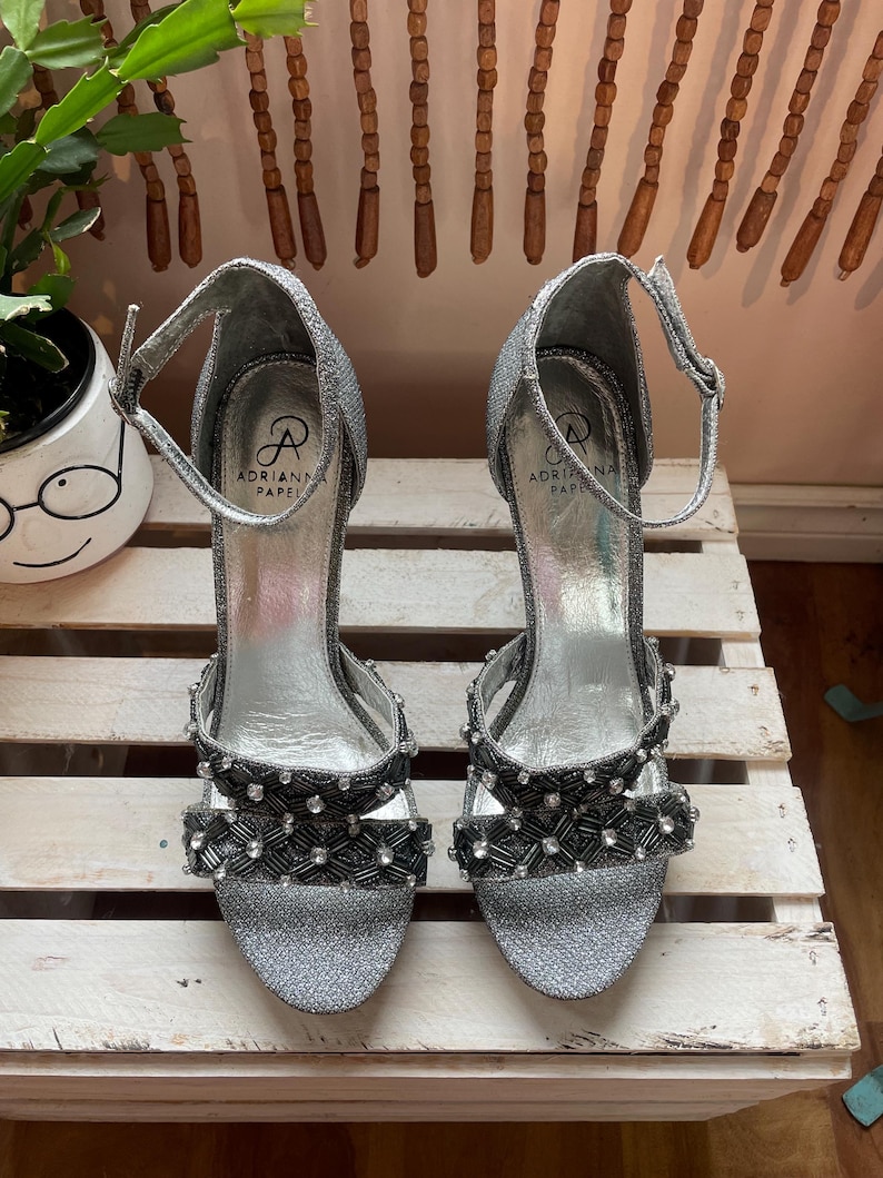 May include: A pair of silver glitter high-heeled sandals with ankle straps. The sandals feature black and silver beaded embellishments on the straps and toe. The brand name "Adrianna Papell" is visible on the insole.