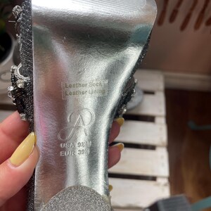 May include: Close-up of a silver high-heeled shoe. The sole is silver with the text "Leather Sock Leather Lining". The shoe size is USA 9 1/2 M, EUR 39 1/2. The shoe has a sparkly, embellished design.
