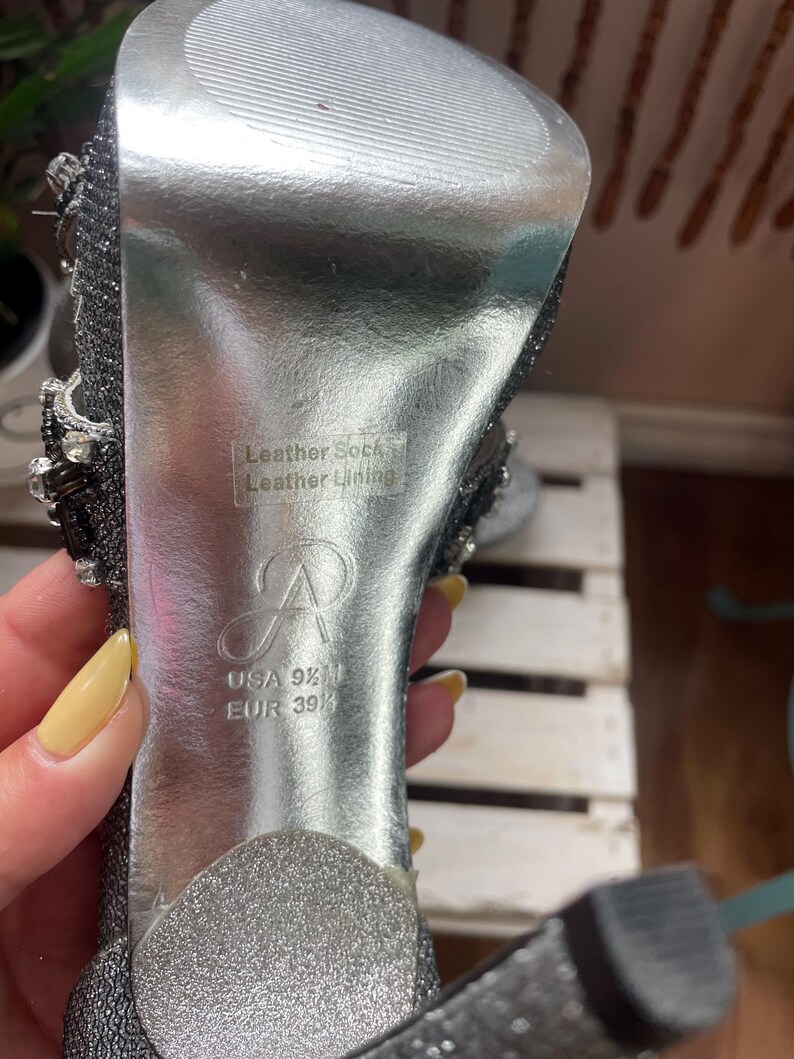 May include: Close-up of a silver high-heeled shoe. The sole is silver with the text "Leather Sock Leather Lining". The shoe size is USA 9 1/2 M, EUR 39 1/2. The shoe has a sparkly, embellished design.