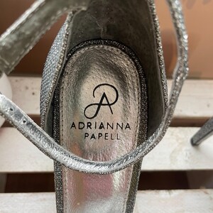 May include: Close-up of a silver high-heeled sandal with a sparkly, glittery texture. The sandal features a thin ankle strap and the brand name "Adrianna Papell" printed on the insole.
