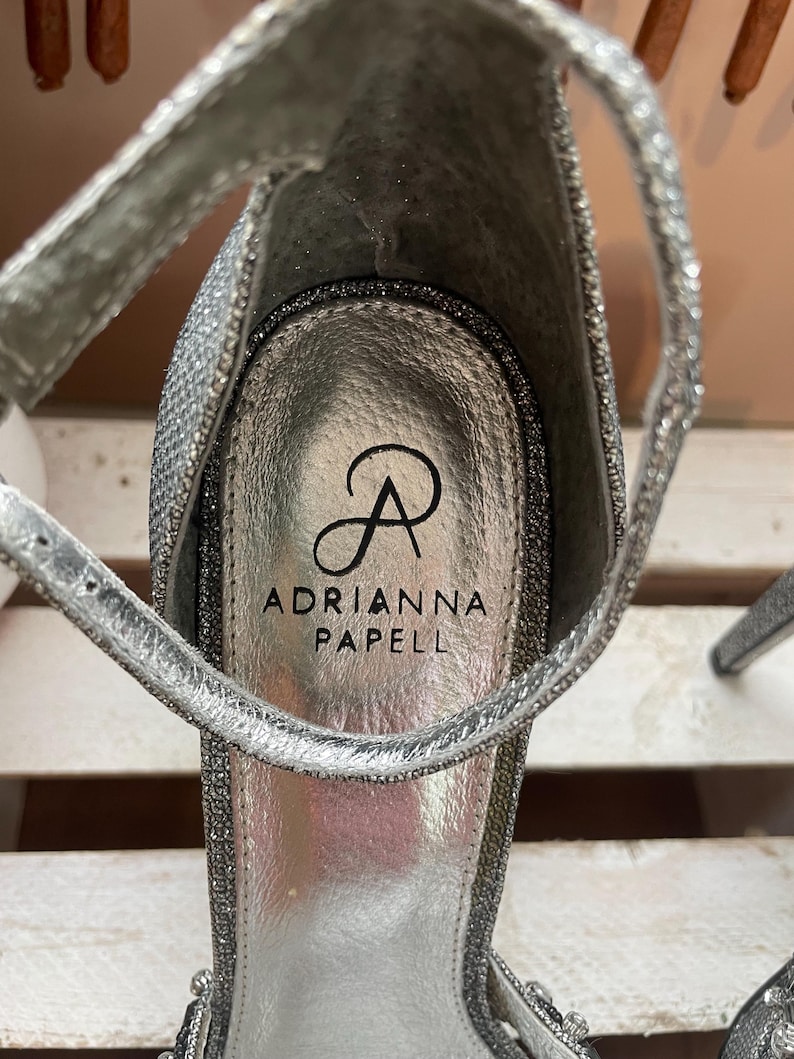 May include: Close-up of a silver high-heeled sandal with a sparkly, glittery texture. The sandal features a thin ankle strap and the brand name "Adrianna Papell" printed on the insole.