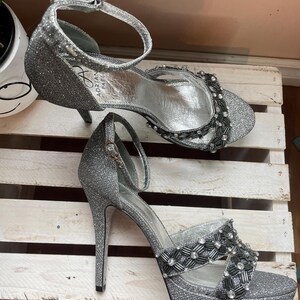 May include: A pair of silver glitter high-heeled sandals. The sandals have a platform sole, ankle straps, and decorative beading across the top. The heels are slender and the inner sole is silver. The brand name "Adrian Papel" is visible on the side.