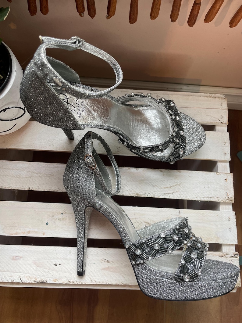 May include: A pair of silver glitter high-heeled sandals. The sandals have a platform sole, ankle straps, and decorative beading across the top. The heels are slender and the inner sole is silver. The brand name "Adrian Papel" is visible on the side.