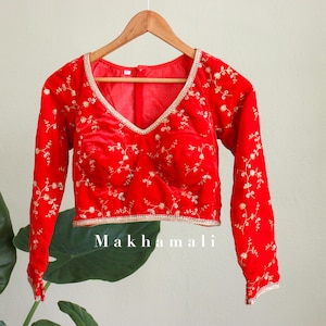 May include: Red velvet blouse with gold embroidery and long sleeves. The blouse has a V-neckline and is cropped. The text "Makhmali" is printed on the bottom of the blouse.