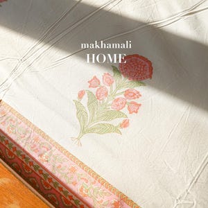 Könnte beinhalten: Weiße Bettwäsche aus Baumwolle mit einem rosa-grünen Blumendruck. Die Bettwäsche hat einen Rand mit einem sich wiederholenden Blumenmuster. Der Text "makhamali HOME" ist auf die Bettwäsche gedruckt.