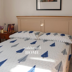 Op de afbeelding: Een wit bed met een blauw-wit gestreept dekbedovertrek en twee bijpassende kussenslopen. Het bed heeft een houten hoofdbord en voetbord. De tekst "makhamali HOME" is op het dekbedovertrek gedrukt.