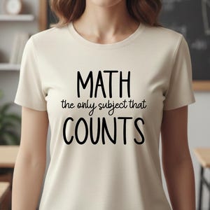 May include: A cream-colored t-shirt with the text "MATH the only subject that COUNTS" in black. The shirt is being worn in a classroom setting, with a chalkboard and desks visible in the background.