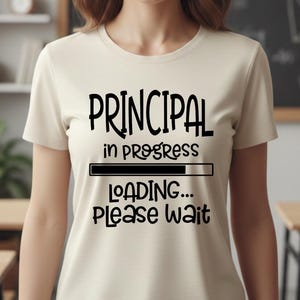 Może przedstawiać: Kobieta w kremowym t-shircie z czarnym napisem "PRINCIPAL in progress LOADING... Please wait". Tło obejmuje tablicę z równaniami, półki na książki i drewniane biurka, sugerując otoczenie szkolne.