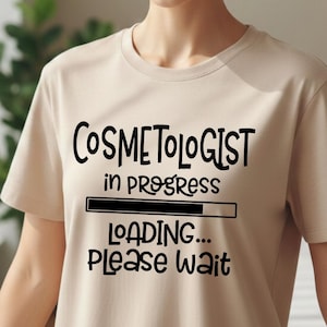 May include: A beige short-sleeved t-shirt featuring black text that reads "COSMETOLOGIST in PROGRESS" above a loading bar graphic, followed by the text "LOADING... PLEASE WAIT" printed in a bold, rounded, sans-serif font on the chest.