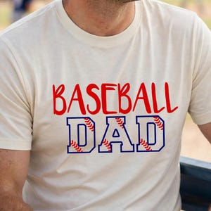 May include: A cream-coloured short-sleeve t-shirt featuring the red text BASEBALL and the blue outlined text DAD with a baseball stitch pattern inside the letters. The shirt has a crew neckline and a standard fit, displaying a graphic design.