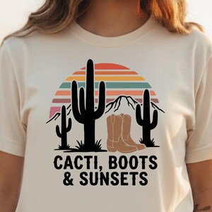 May include: Cream-colored t-shirt featuring a graphic design with cacti, cowboy boots, and a sunset. The text "CACTI, BOOTS & SUNSETS" is printed below the graphic. The shirt is worn in a field of sunflowers.