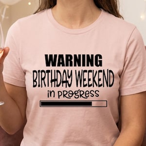 May include: A light pink t-shirt with the text "WARNING BIRTHDAY WEEKEND in PROGRESS" in black. A loading bar graphic is below the text. A person is holding a glass of wine.