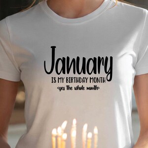 May include: White t-shirt with the text "January is my birthday month - yes the whole month" in black script. The shirt is being held in front of a cake with lit candles.