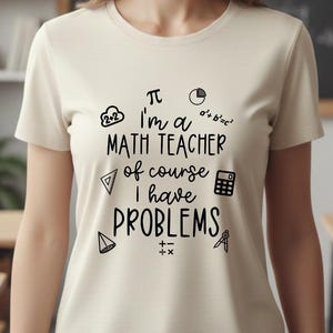 May include: A cream-colored short-sleeve t-shirt featuring black text that reads "I'm a MATH TEACHER of course I have PROBLEMS" surrounded by various mathematical symbols and icons including a calculator, a pi symbol, and geometric shapes.
