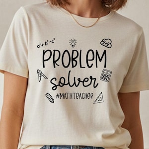 May include: A cream-coloured t-shirt featuring the words "PROBLEM solver" in black script, accompanied by math symbols and the hashtag #MATHTEACHER. The shirt is worn with blue jeans.