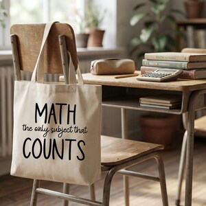 May include: A beige canvas tote bag with the text "MATH the only subject that COUNTS" in black. The bag hangs on the back of a wooden school chair. A vintage school desk with books and a calculator is in the background.