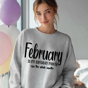 May include: A light gray sweatshirt with the word "February" in large black script. Below, the text reads "IS MY BIRTHDAY MONTH" and "yes the whole month" in a smaller font. The image includes balloons in the background.