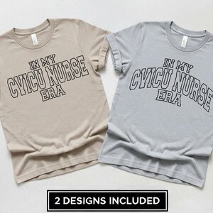 以下が含まれることがあります： ベージュとヘザーグレーの半袖Tシャツ2枚。それぞれに「IN MY CVICU NURSE ERA」という黒いカレッジスタイルの文字がプリントされています。Tシャツはクルーネックのスタンダードなフィット感で、下部には黒いラベルが付いています。