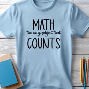 May include: Light blue t-shirt with the text "MATH the only subject that COUNTS" in black. The shirt is displayed on a light wood surface, with a stack of books and pencils nearby. A casual, everyday wear tee.