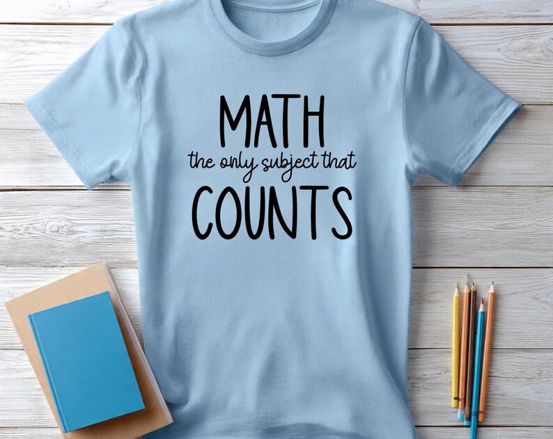 May include: Light blue t-shirt with the text "MATH the only subject that COUNTS" in black. The shirt is displayed on a light wood surface, with a stack of books and pencils nearby. A casual, everyday wear tee.