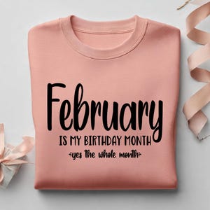 May include: A peach-colored sweatshirt with the words "February IS MY BIRTHDAY MONTH" in black script. The phrase "yes the whole month" is written below. A gift box and ribbon are in the background.