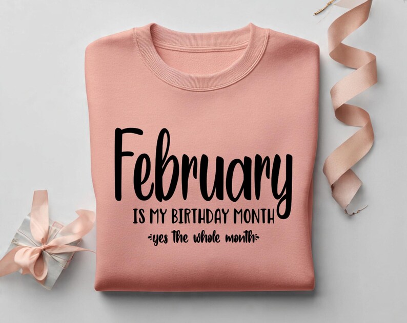 May include: A peach-colored sweatshirt with the words "February IS MY BIRTHDAY MONTH" in black script. The phrase "yes the whole month" is written below. A gift box and ribbon are in the background.