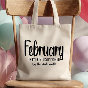 May include: A cream-colored tote bag with the text "February is my birthday month - yes the whole month" in black script. The bag is on a wooden chair, with colorful balloons in the background.