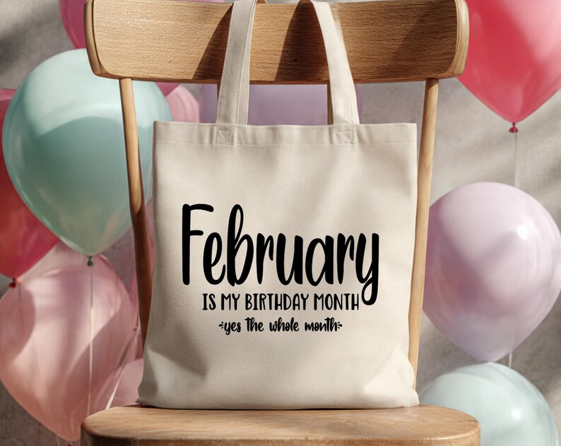 May include: A cream-colored tote bag with the text "February is my birthday month - yes the whole month" in black script. The bag is on a wooden chair, with colorful balloons in the background.