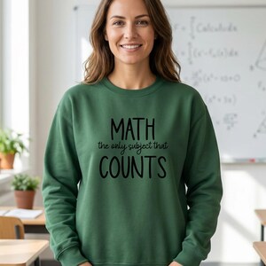 May include: A forest green long-sleeve crewneck sweatshirt featuring the black text "MATH the only subject that COUNTS" printed on the front. The garment has a ribbed neckline, cuffs, and hem, providing a standard fit for everyday wear.