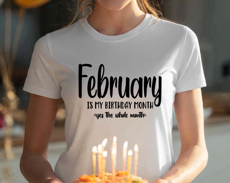 May include: White t-shirt with the text "February is my birthday month - yes the whole month" in black script. The person is holding a birthday cake with lit candles. The shirt is a crew neck.