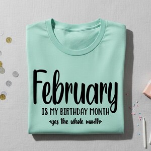 May include: A light green t-shirt with the text "February IS MY BIRTHDAY MONTH - yes the whole month" in black script. The shirt is folded neatly on a light gray surface, with confetti and a small gift box nearby.