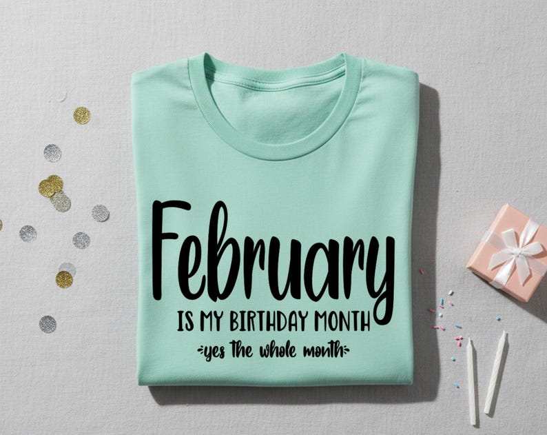 May include: A light green t-shirt with the text "February IS MY BIRTHDAY MONTH - yes the whole month" in black script. The shirt is folded neatly on a light gray surface, with confetti and a small gift box nearby.