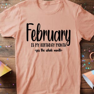 May include: A peach-colored t-shirt with the text "February IS MY BIRTHDAY MONTH - yes the whole month" in black script. The shirt is displayed on a wooden surface with party decorations.