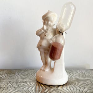 May include: White porcelain figurine of a person holding a small hourglass. The hourglass is filled with brown sand and has a clear glass bulb.