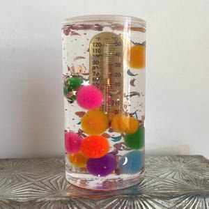 Op de afbeelding: Een heldere, cilindrische vloeistofthermometer met een goudkleurige temperatuurmeter. De thermometer is gevuld met kleurrijke pompons en glitter. De temperatuurschaal is gemarkeerd in Fahrenheit en Celsius.