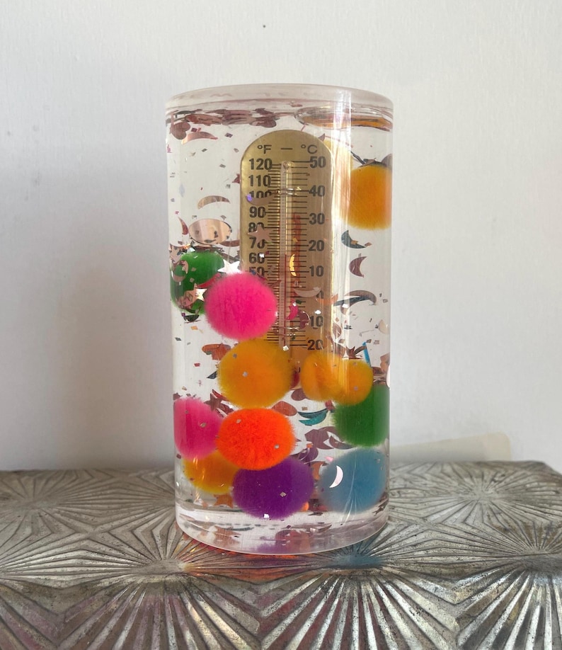 Op de afbeelding: Een heldere, cilindrische vloeistofthermometer met een goudkleurige temperatuurmeter. De thermometer is gevuld met kleurrijke pompons en glitter. De temperatuurschaal is gemarkeerd in Fahrenheit en Celsius.